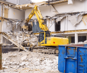 Yellow excavator tearing down a building with a blue roll-off in the foreground. 