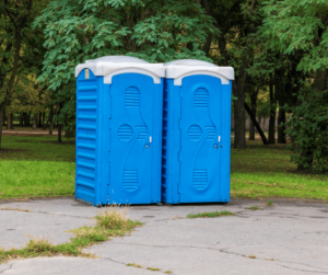 Pair of blue portable restrooms on a concrete slab in front of trees.